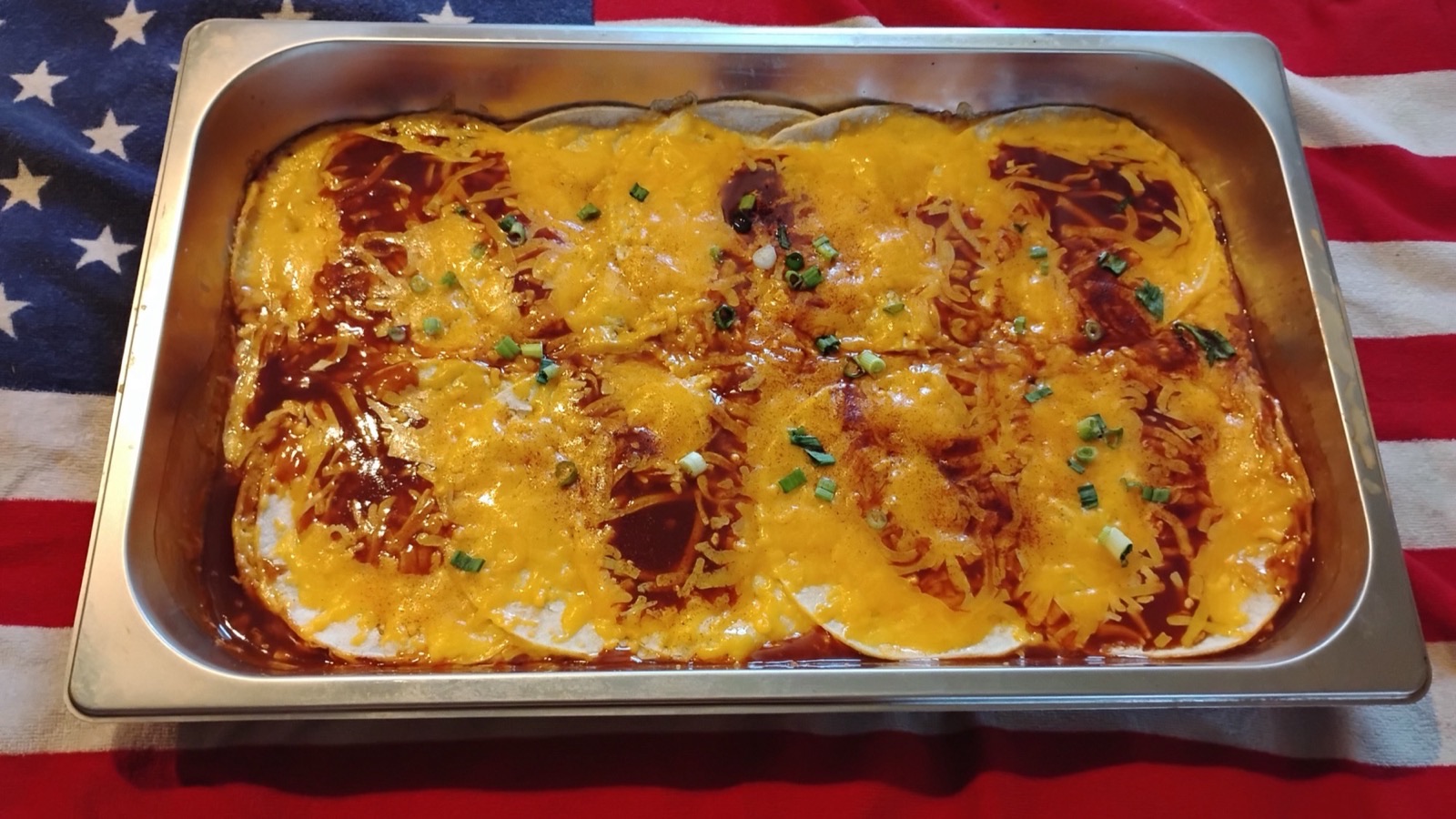 A catering tray of cheesy enchiladas topped with red sauce, melted cheddar and green onion